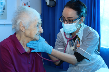 Picture of a nurse helping a patient. The nurse wears a face mask and glasses