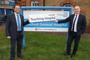 Picture of staff smiling and standing in front of the Watford Hospital sign post holding a card with with the words 'teaching hospital' written on it
