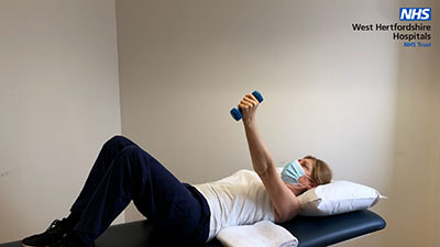 Picture of a person lying on a bed, holding a weight raised in their left hand